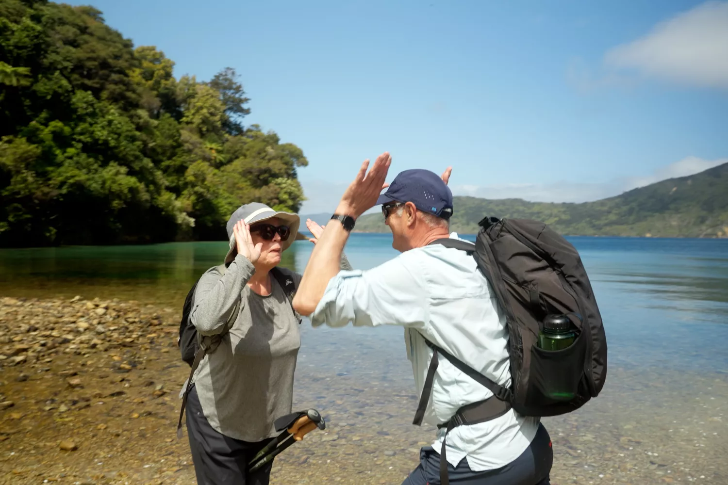 Two individuals with backpacks stand on the shore of a serene lake high-fiving, surrounded by nature's beauty.